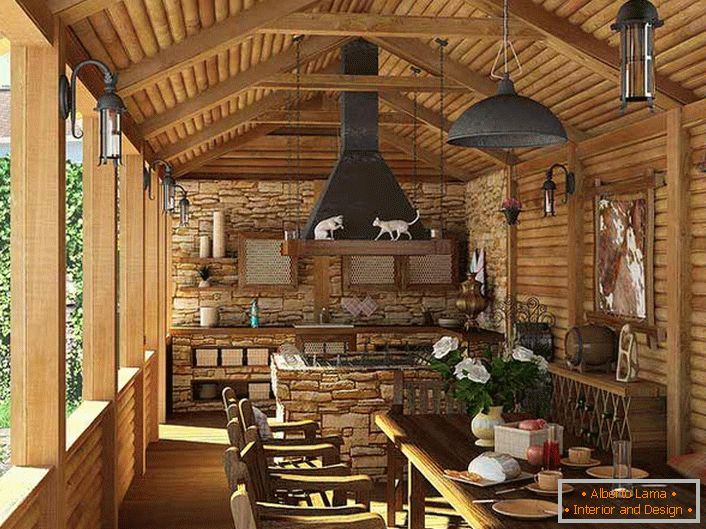 Uma pequena cozinha com churrasqueira na varanda de uma casa de campo. O estilo do país é evidenciado, em primeiro lugar, pela decoração das paredes e do teto com uma moldura de madeira.