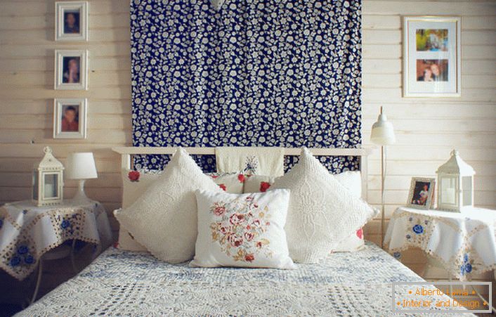 De acordo com o estilo rústico, a cama é decorada com vários travesseiros com bordados vermelhos contrastantes. Mesas de cabeceira são cobertas com uma toalha de mesa com delicadas flores azuis.